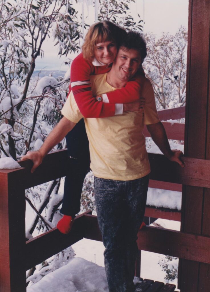 A smiling couple on a snowy wooden balcony, she hugging him from behind