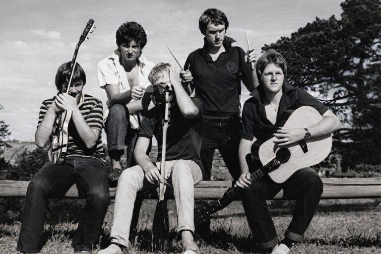 Black and white photo of The Innocent Bystanders, five young men with guitars and drumsticks on a park bench